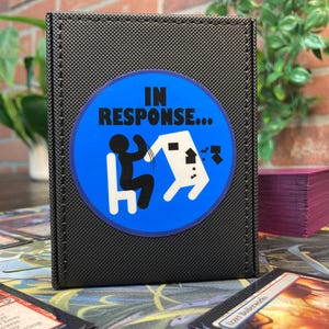 May include: Black card storage box with a blue circular graphic that reads "IN RESPONSE..." featuring a stick figure and a white shape. The box is textured and has a stitched border. A stack of purple cards is visible in the background.