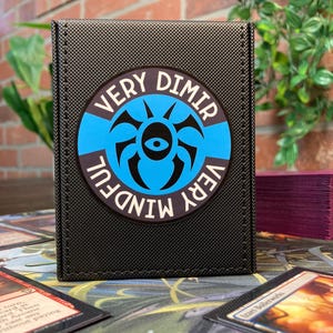 May include: Black card storage box with a blue and white circular emblem that reads "VERY DIMIR VERY MINDFUL". The box is sitting on a table with playing cards.