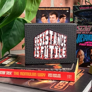 May include: Black wallet with the phrase "Resistance is Futile" in white and red lettering. The wallet is displayed on top of several Star Trek books, including "The Romulan Way."