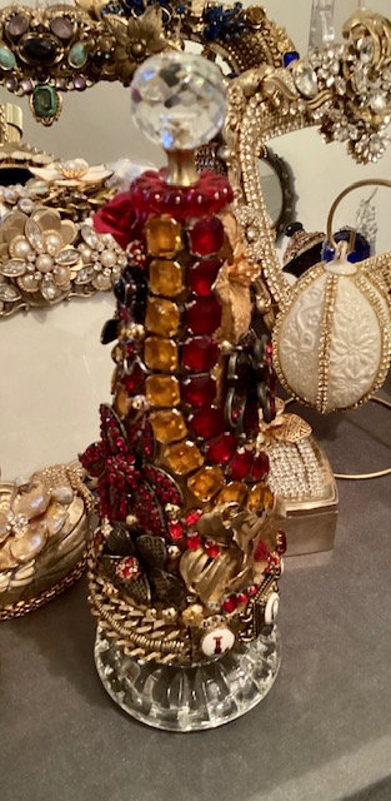May include: A decorative bottle stopper made of glass, rhinestones, and metal. The stopper is shaped like a Christmas tree and is decorated with red, gold, and clear rhinestones. The stopper is topped with a clear crystal.