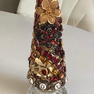 May include: A decorative glass bottle with a gold top and a cone-shaped body covered in red, gold, and brown jewels. The bottle is decorated with a gold flower and the word "LOVE" in gold letters.