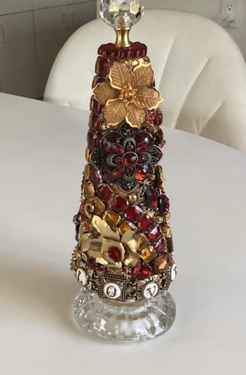 May include: A decorative glass bottle with a gold top and a cone-shaped body covered in red, gold, and brown jewels. The bottle is decorated with a gold flower and the word "LOVE" in gold letters.