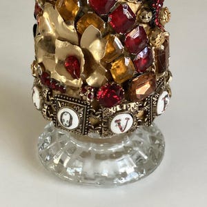 May include: A glass bottle with a gold and red jeweled top. The bottle has a clear glass base and the top is decorated with rhinestones, beads, and metal accents. The word "LOVE" is spelled out in white letters on the bottle.
