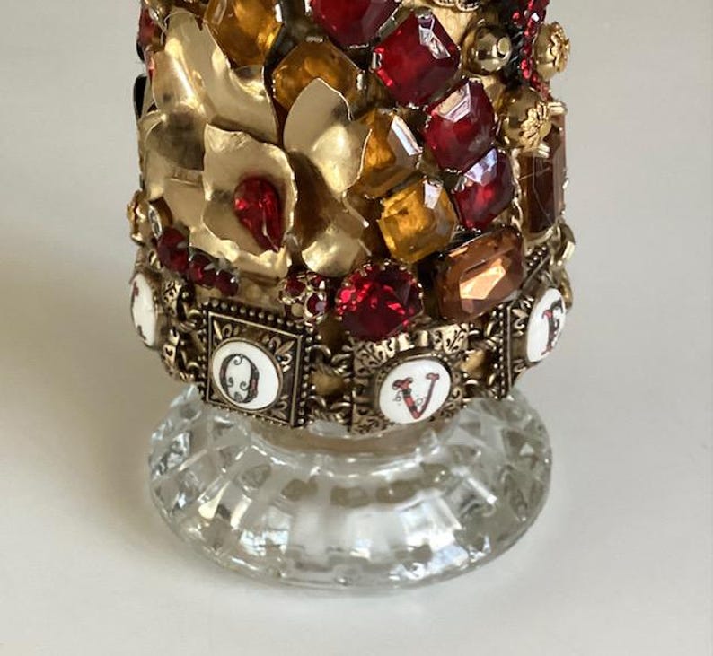 May include: A glass bottle with a gold and red jeweled top. The bottle has a clear glass base and the top is decorated with rhinestones, beads, and metal accents. The word "LOVE" is spelled out in white letters on the bottle.
