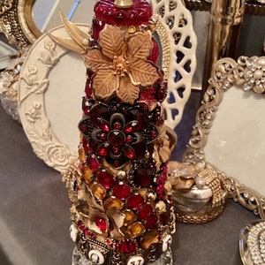 May include: A cone-shaped Christmas tree ornament decorated with red and gold jewels, rhinestones, and a gold flower. The ornament is topped with a clear crystal ball.