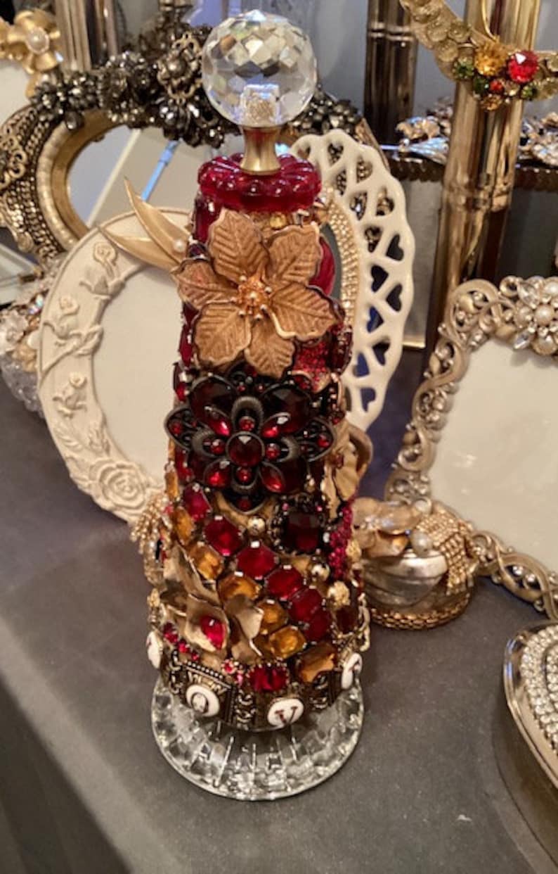 May include: A cone-shaped Christmas tree ornament decorated with red and gold jewels, rhinestones, and a gold flower. The ornament is topped with a clear crystal ball.