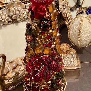 May include: A decorative bottle stopper made of glass and rhinestones. The bottle stopper is shaped like a cone and is covered in red, gold, and black rhinestones. The bottle stopper is sitting on a clear glass base.