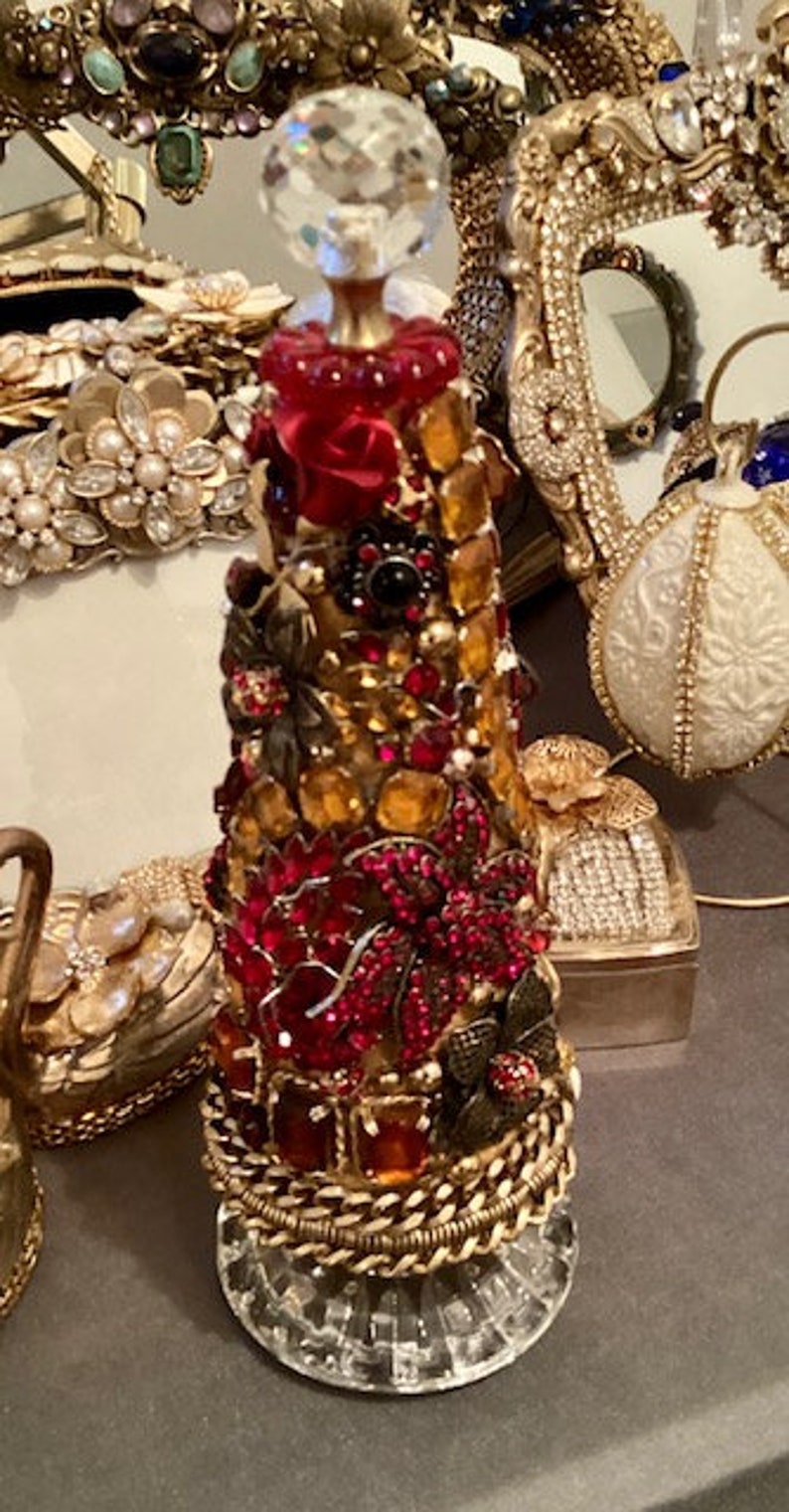 May include: A decorative bottle stopper made of glass and rhinestones. The bottle stopper is shaped like a cone and is covered in red, gold, and black rhinestones. The bottle stopper is sitting on a clear glass base.