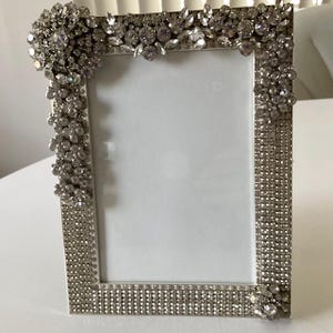 May include: Silver rectangular picture frame with a white interior. The frame is heavily embellished with sparkling rhinestones and crystals, creating a glamorous and ornate design. The corners and top edge feature clusters of larger stones.