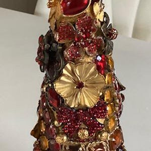 May include: A decorative bottle stopper made from a variety of materials, including glass, metal, and jewels. The bottle stopper is shaped like a cone and is decorated with red, gold, and clear jewels. The bottle stopper is sitting on a clear glass base.