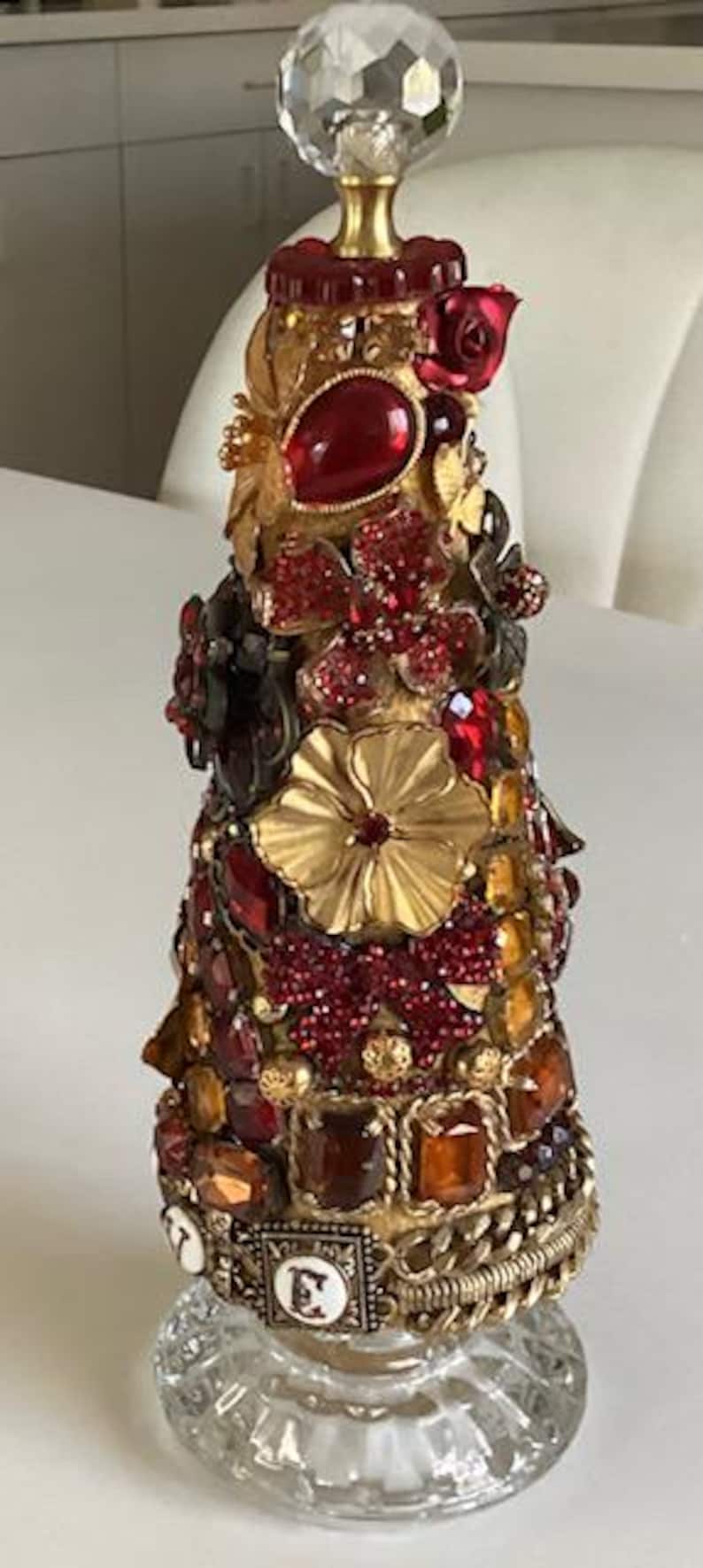 May include: A decorative bottle stopper made from a variety of materials, including glass, metal, and jewels. The bottle stopper is shaped like a cone and is decorated with red, gold, and clear jewels. The bottle stopper is sitting on a clear glass base.