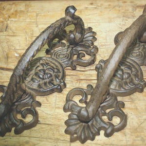 May include: Two ornate, cast iron drawer pulls with a rustic, antique finish. Each pull features a curved handle and decorative base with floral and scroll details. The hardware is a dark, weathered brown.