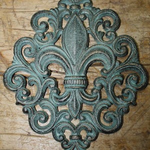 May include: A cast iron wall decor piece in a fleur-de-lis design. The decorative item features a distressed teal and black finish with intricate scrollwork and a central spearhead shape. The piece is mounted on a wooden surface.