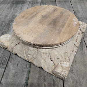 May include: A rustic, square-based wooden pedestal with a circular top. The pedestal is painted in a distressed, off-white color, with carved leaf-like details on the sides. The top is a natural wood color.