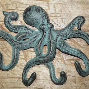 May include: A cast iron octopus wall hook with a distressed teal and black finish. The octopus has eight curved tentacles that serve as hooks. The decorative nautical-themed piece is mounted on a wooden surface.