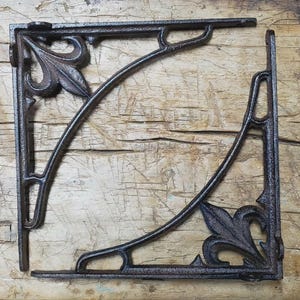 May include: Two ornate, dark brown cast iron shelf brackets. Each bracket features a decorative leaf design at the corner and a curved support arm. The brackets are designed to be mounted on a wall to support a shelf.