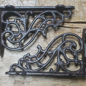 May include: Two ornate, cast iron shelf brackets. The brackets are dark brown with a distressed finish, featuring a scrolling, openwork design. The brackets are L-shaped, with mounting holes for easy installation. Perfect for adding a touch of vintage charm to any space.