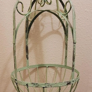 May include: A hanging metal basket with a distressed, light green finish. The basket has a rounded bottom and a decorative, scrolled design. The metal shows signs of wear, giving it a vintage look. The basket is suspended by a chain.