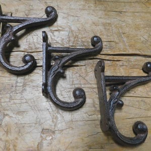 May include: Three ornate, cast iron wall hooks with a rustic, brown finish. Each hook features a decorative scroll design and a rounded end. The hooks are mounted on a wooden surface, suggesting a vintage or farmhouse aesthetic.
