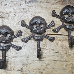 May include: Three cast iron skull and crossbones wall hooks. The hooks are dark brown with a textured finish, and each features a skull with crossed bones and a single hook below. They are mounted on a weathered wooden surface.