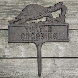 May include: A cast iron garden stake with a turtle design and the words "TURTLE CROSSING" in raised letters. The stake is brown and has a textured surface, suitable for outdoor use. The turtle is depicted crawling towards some plants.