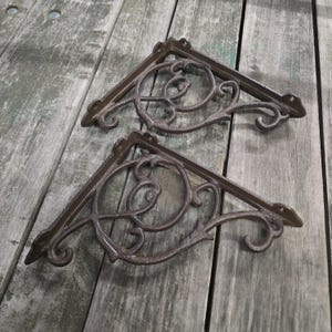 May include: Two dark brown metal shelf brackets featuring a triangular frame with an intricate, curved scrollwork design. Each bracket has mounting holes at the corners and a textured finish, providing a sturdy support structure for shelving.