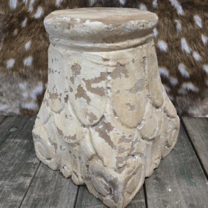 May include: A weathered, beige-colored pedestal with a round top and a flared, textured base. The base features a repeating, stylized leaf pattern. The pedestal has a distressed, aged appearance, suggesting it is an antique or vintage item.