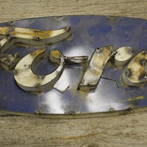 May include: A vintage Ford sign with a distressed, weathered appearance. The oval-shaped sign features the word "Ford" in a stylized font, with a blue background and metallic lettering. The sign is mounted on a wooden surface.