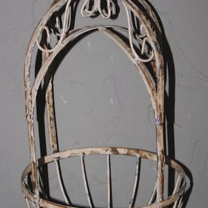 May include: Off-white, ornate metal hanging basket with a distressed finish. The basket features a rounded base and a decorative top with scrollwork and a chain for hanging. Ideal for displaying plants or decor.