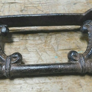 May include: A dark brown, cast iron towel bar with an ornate design. The bar has a horizontal rod with rounded ends and is supported by curved brackets with decorative flourishes. The mounting plate has two holes.