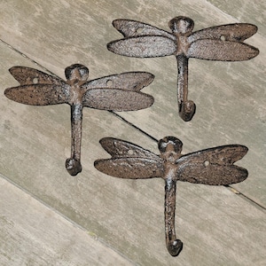 May include: Three cast iron dragonfly hooks with a rustic brown finish. Each hook features a dragonfly design with detailed wings and a curved hook for hanging items. The hooks are mounted on a wooden surface.