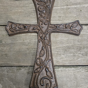May include: A decorative, cast iron cross with an ornate, swirling leaf and vine design. The cross is a dark brown color and has a hole at the top for hanging. The background is a weathered wooden surface.