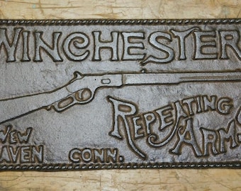 May include: A rectangular, cast iron sign with the words "WINCHESTER REPEATING ARMS" and a rifle silhouette. The sign also includes "New Haven, Conn." in a decorative font. The sign has a rope-like border.