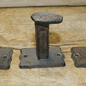 May include: Three rustic, cast iron nail head hooks. Each hook has a rectangular base with two holes for mounting and a nail head shaped top. The hooks are a dark, weathered brown color.