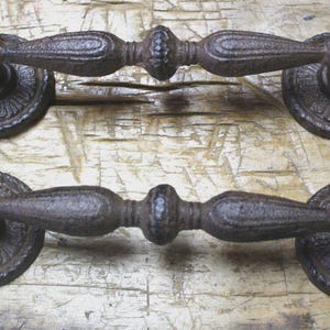 May include: Two antique-style, cast iron door handles. The handles are dark brown with ornate detailing, including a central bulbous section and decorative circular backplates. The handles are displayed on a weathered wooden surface.