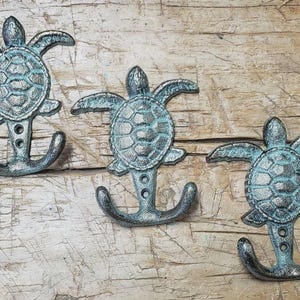 May include: Three cast iron wall hooks in the shape of sea turtles. The hooks are painted in a distressed teal and silver finish, with an anchor-shaped base. The hooks are mounted on a wooden surface.