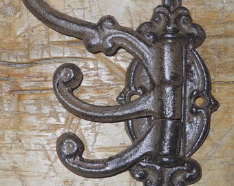 Può includere: Un gancio da parete decorativo in ghisa con tre bracci curvi. Il gancio presenta un design ornato con una finitura scura e usurata. La piastra di montaggio è di forma ovale con decorazioni decorative.