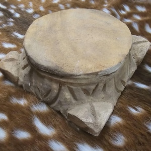 May include: A beige, carved pedestal with a round top. The pedestal has a decorative, geometric design around the base. The object is made of a light-colored material and sits on a brown and white patterned surface.