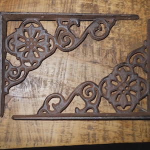 May include: Two ornate, cast iron shelf brackets with a rust-colored finish. Each bracket features a floral design at the corner and decorative scrollwork. The brackets are designed to be mounted at a 90-degree angle.