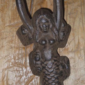 May include: A dark brown cast iron mermaid wall hook. The mermaid has her arms raised above her head. The mermaid's tail is curved. The hook is mounted on a light brown wooden surface.