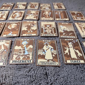 May include: A set of tarot cards with pixelated illustrations in brown and beige tones. Each card displays a distinct design and title, such as "Star," "Moon," and "Lovers." The cards are laid out on a grey surface.