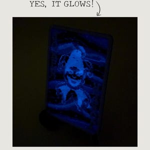 Puede incluir: Imagen azul brillante de la cara de un payaso en un objeto rectangular. El texto "YES, IT GLOWS!" está encima de la imagen. La cara del payaso está iluminada con una luz azul brillante.