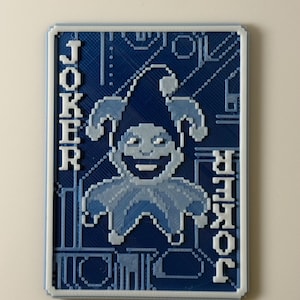 Puede incluir: Una carta de juego Joker impresa en 3D, azul y blanca. La carta presenta un diseño de bufón pixelado, con la palabra «JOKER» impresa verticalmente en ambos lados. El diseño es en tonos azules y blancos.