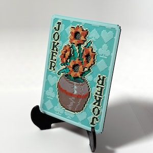 Peut inclure: Une plaque rectangulaire sur fond bleu clair présentant un motif en pixel art de tournesols orange dans un pot marron. Le mot JOKER est imprimé verticalement sur les côtés gauche et droit. Le fond comporte un motif répété de symboles de cartes.
