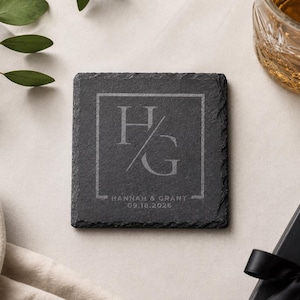 May include: A square black slate coaster featuring a laser-engraved design with the letters H and G separated by a diagonal line inside a square border. Below the letters, the text reads HANNAH & GRANT and the date 09.18.2026 in white lettering.