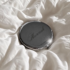 May include: A round, silver-toned metal compact mirror with a polished, reflective surface. The name Hannah is engraved in a cursive script across the center of the lid. A small hinge is visible on the side, and a bright light reflection sparkles.