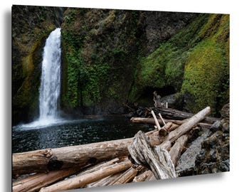 Könnte beinhalten: Ein Foto eines Wasserfalls, der in einen dunklen Wasserpool stürzt, umgeben von grüner Vegetation und moosbewachsenen Felsen. Verwitterte Baumstämme im Vordergrund bilden eine natürliche Szene.