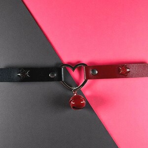 May include: A black and red choker necklace with a heart-shaped pendant and a small red bell. The choker has star-shaped studs and a silver heart-shaped connector. The background is divided diagonally into gray and pink sections.