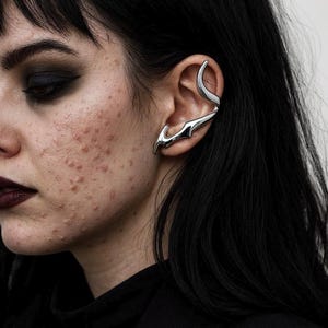 May include: A silver ear cuff with a curved design, worn on the ear. The ear cuff has a sleek, modern aesthetic. The jewelry piece curves around the ear and features a pointed, flame-like design.