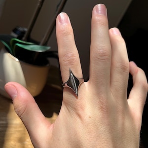 May include: A silver-colored ring with a geometric, diamond-shaped design. The ring is worn on a finger, showcasing its polished surface and modern aesthetic. The ring's design features a central, dark-colored inlay.